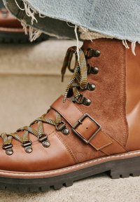 Braune Leder- und Wildleder-Wanderschuhe mit mehrfarbigen Schnürsenkeln, metallischen Ösen, einem geschnallten Riemen und einer strukturierten Gummisohle.