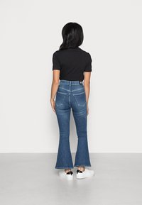 Högmidjade denimutställda jeans i mediumblått med en slät textur. Har två bakfickor och en etikett på midjebandet. Åtföljs av vita sneakers.