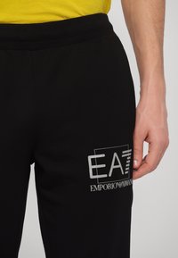 Black sweatpants made of soft material, featuring an elastic waistband and a white logo print of "EA7 Emporio Armani" on the left thigh.
