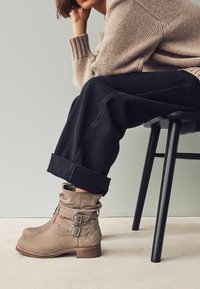Botines de cuero beige con un diseño holgado, acentos de doble hebilla y textura acolchada, combinados con pantalones de mezclilla oscuros remangados.