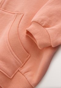 Virvendav roosa pusa lähivaade, kus on näha soonikkoes varruka- ja vöökohaääred ning eesmine kängurutasku õmblused.