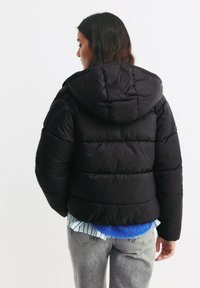 Zwarte pufferjack met een capuchon, gestikte textuur, elastische manchetten en een cropped snit. Draag over een meerkleurig gelaagd shirt en grijze jeans.