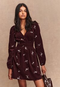 Vestito in velluto a coste color borgogna con maniche lunghe, scollo a V, ricamo floreale e vita increspata. Abbinato a una borsa marrone.