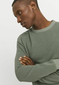 Grüner Strickpullover mit strukturiertem Muster, geripptem Halsausschnitt und Bündchen, präsentiert an einer Person mit verschränkten Armen.