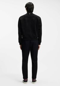 Black suede jacket with ribbed cuffs and hem, featuring a stand collar and subtle stitching details, worn with dark trousers and shoes.