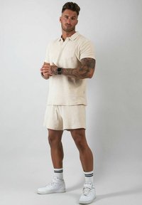 Beige bomullspolo med kort ärm och bröstficka, kombinerad med matchande shorts. Bärs med vita sneakers och randiga svartvita strumpor.