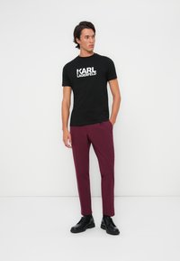 Černé tričko s bílým logem "KARL LAGERFELD", zkombinované s vínovými kalhotami a černými šněrovacími botami. Minimalistický design, hladká textura.