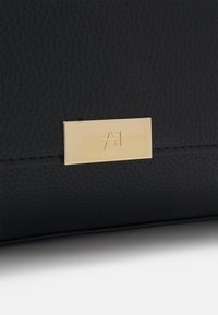 Sac à main en cuir noir avec une surface texturée. Il comprend une plaque logo dorée avec des initiales embossées et un design élégant.
