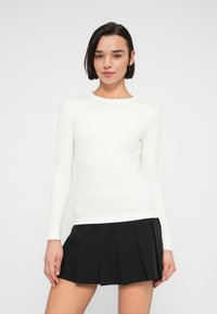 Λευκό εφαρμοστό τοπ με μακριά μανίκια και στρογγυλή λαιμόκοψη, σε συνδυασμό με μαύρη πλισέ φούστα. Ομαλή υφή, μινιμαλιστικός σχεδιασμός.