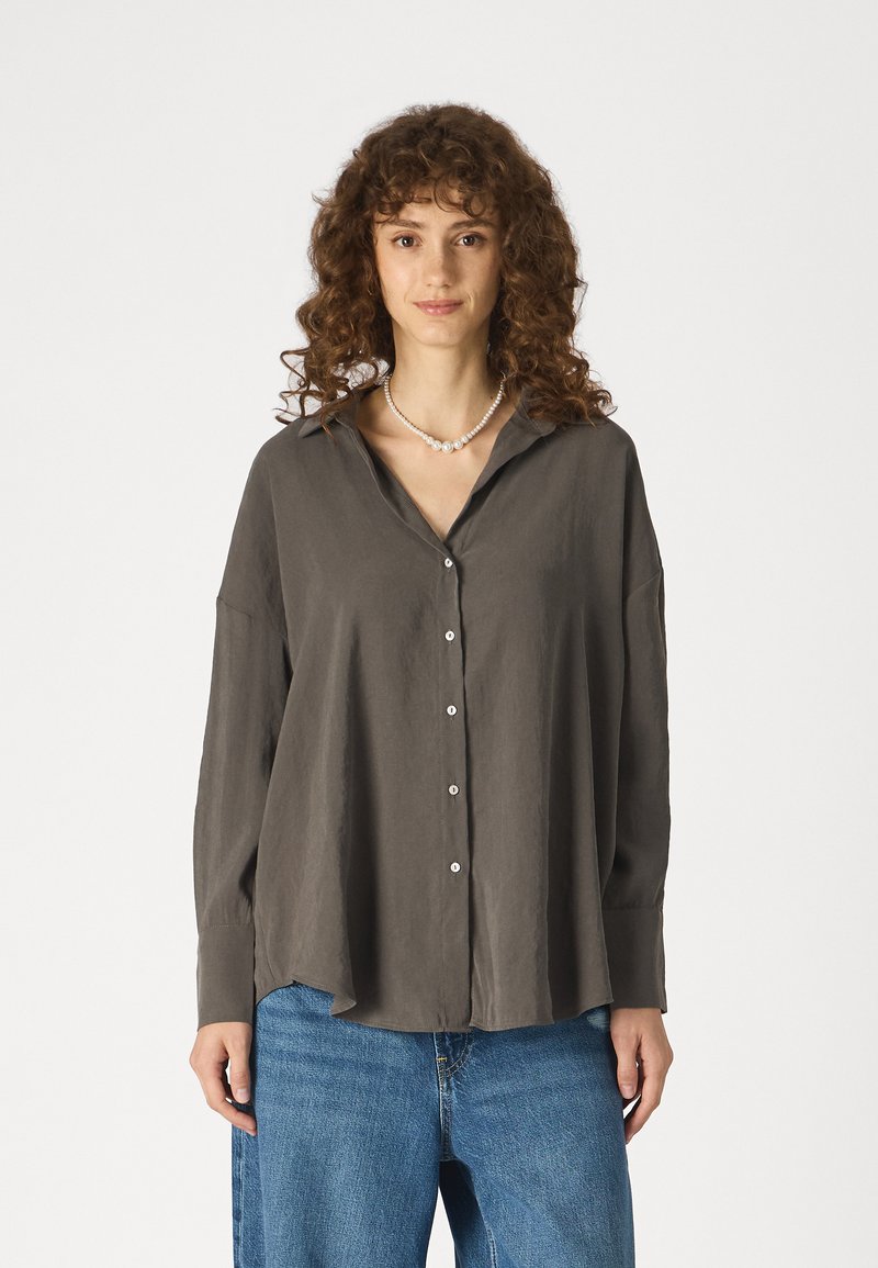 Camisa cinza escura, de botões, com um corte solto, mangas longas e colarinho. Feita de um tecido leve. Usadas com um par de jeans azuis largos.