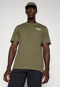 Camiseta de manga corta en verde oliva, hecha de una tela suave, que presenta un pequeño logo impreso en el área del pecho izquierdo. Diseño simple y recto.