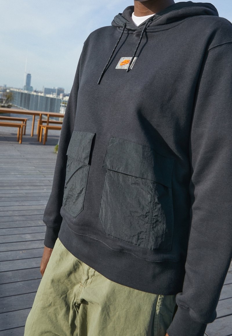 Svart hoodie med justerbar dragsko, med två stora matte svarta fickor framtill. Bärs över ljusgröna cargobyxor.