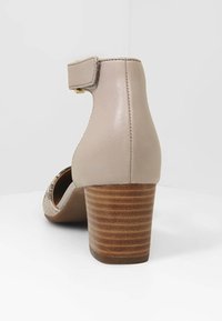 Beige nahka korko, jossa on pyöreä muoto ja puupanelista valmistettu pohja. Varustettu säädettävällä nilkkahihnalla ja minimalistisella designilla.