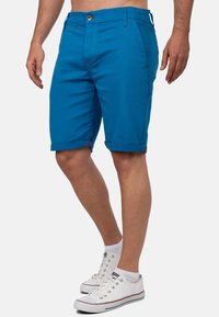 Blauwe katoenen shorts met opgerolde zomen, rode stiksels en knoopsluiting. Draagt met witte sneakers en zichtbare enkels.