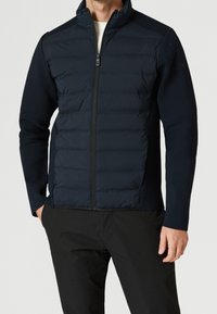 Chaqueta azul marino con cremallera frontal, cuerpo acolchado, mangas de punto y cuello alto. Textura suave con detalle de costuras horizontales.