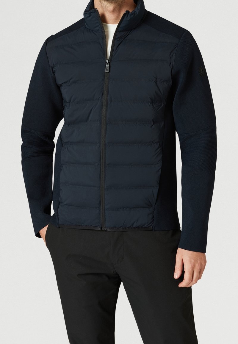 Chaqueta azul marino con cremallera frontal, cuerpo acolchado, mangas de punto y cuello alto. Textura suave con detalle de costuras horizontales.