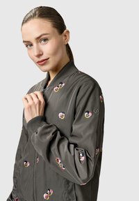 Ung kvinde med trukket hår tilbage, iført en grå bomberjakke med pink og gul blomsterbroderi, der holder i lynlåsen nær kraven.