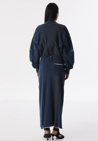 Lang, getailleerde jurk van donkerblauwe stof met een mix van texturen. Heeft ballonmouwen, een split aan de achterkant en praktische zijzakken.