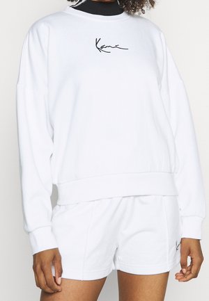 Osoba nosící bílou mikinu s dlouhými rukávy a odpovídající šortky s malými černými kurzívními logy na obou oděvech.