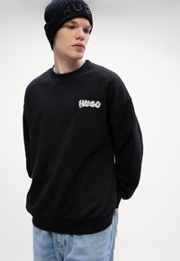 Felpa nera in tessuto strutturato con orlo a coste, caratterizzata da un logo "HUGO" ricamato in bianco. Indossata con un cappello di lana abbinato.