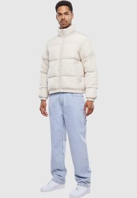 Jachetă tip puffer de culoare bej deschis, cu secțiuni quilted orizontale și guler înalt. Asortată cu pantaloni din denim albastru deschis și adidași albi.