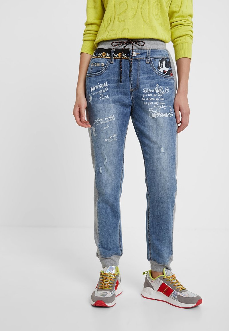 Personne portant un jean bleu avec des motifs manuscrits blancs, un pull jaune et des baskets grises avec des détails rouges et jaunes, debout.