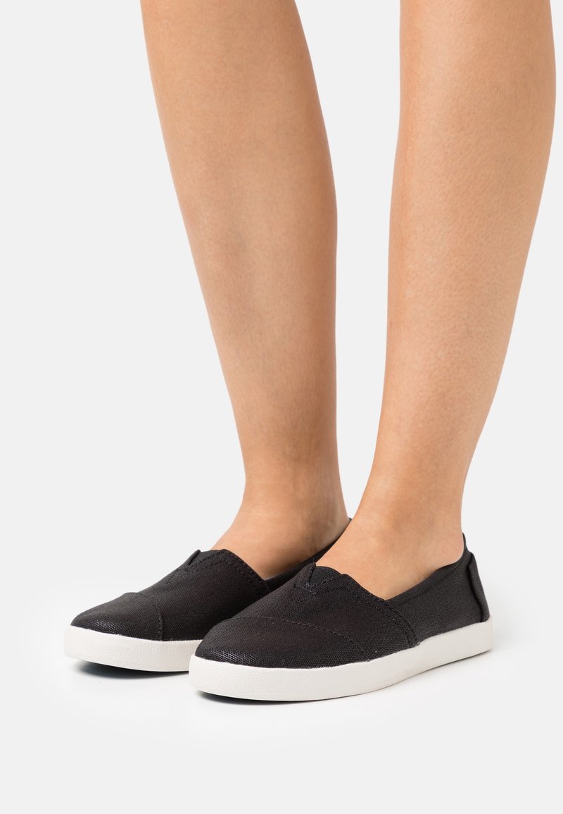 Jambes portant des chaussures en toile noires à enfiler avec semelles en caoutchouc blanches sur un fond blanc.