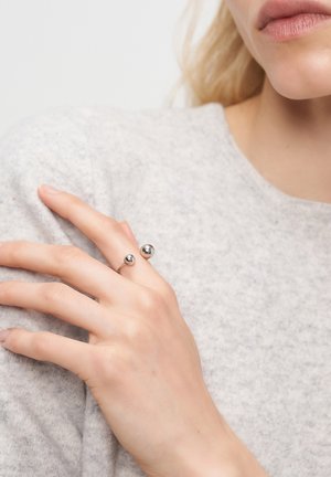 Metallring mit zwei kugelförmigen Akzenten. Silberfarbe, glatte Oberfläche, getragen an einer Hand, die auf einem strukturierten grauen Pullover ruht.