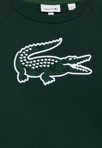 Sudadera de color verde oscuro con un logo bordado de cocodrilo blanco que presenta un patrón geométrico en verde oscuro en su cuerpo.