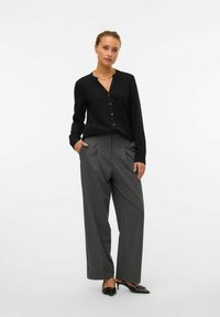 Blouse noire à manches longues avec un col en V et des détails de boutons, assortie à un pantalon plissé gris taille haute. Élégantes chaussures à talons noirs.