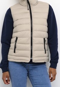 Beige gewatteerd vest met hoge kraag, ritssluiting en zijzakken. Voorzien van een doorgestikt ontwerp en zwarte metalen accenten. Gecombineerd met een donkermarineblauwe trui.