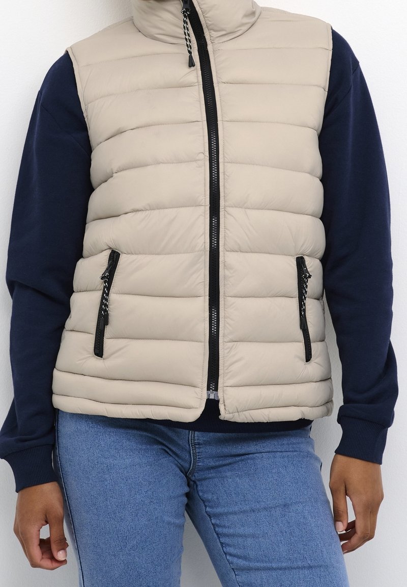 Beige gewatteerd vest met hoge kraag, ritssluiting en zijzakken. Voorzien van een doorgestikt ontwerp en zwarte metalen accenten. Gecombineerd met een donkermarineblauwe trui.