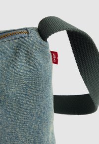 Primo piano dell'angolo di una borsa in denim blu con zip in ottone, etichetta in tessuto rosso e anello del manico intrecciato verde scuro su uno sfondo chiaro.