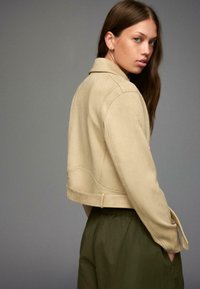 Beige kort skinnjacka i mocka med krage, långa ärmar och sidobälte, matchad med olivgröna löst sittande byxor.