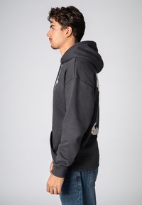 Donkergrijze hoodie met een voorzak, geribbelde mouwen en een verstelbare trekkoord. Voorzien van grafische prints op de mouwen. Soepele textuur.