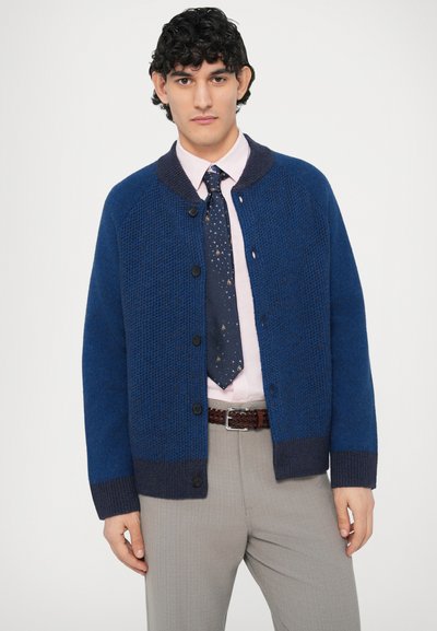 Blauer Strickcardigan mit strukturiertem Muster, kontrastierendem dunkelblauem Kragen, Knöpfen vorne und gerippten Bündchen. Getragen über einem rosa Hemd und Krawatte.