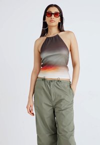 Mulher vestindo um top halter com gradiente, calças verdes soltas, grandes brincos de argola e óculos de sol com lentes avermelhadas, em pé com uma mão no bolso.