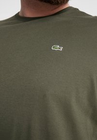 Uomo che indossa una maglietta verde oliva con un piccolo logo di coccodrillo ricamato di verde sul petto.