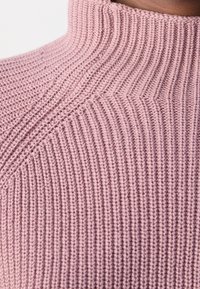 Gros plan sur un pull à col roulé tricoté rose montrant une texture côtelée et un motif de points diagonal sur le col et la poitrine.