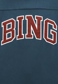 Σκούρο τιρκουάζ ύφασμα με μεγάλα, έντονα κόκκινα γράμματα περιγραμμένα με λευκό που σχηματίζουν τη λέξη "BING" κεντημένα στην επιφάνεια.