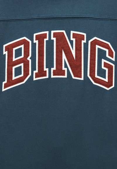 Mørkt blågrønt stoff med store, fete, røde bokstaver med hvit kant som staver "BING" sydd over flaten.