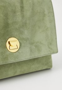 Sac à main en daim vert avec un fermoir circulaire en ton or et des lignes épurées, doté d'un détail de bord cousu sur le rabat.