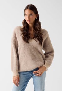 Lichtbeige gebreide trui met een V-hals, gestructureerde stof, brede mouwen en geribbelde manchetten, gecombineerd met lichtblauwe spijkerbroek.