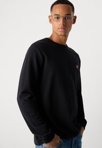 Ung mand iført klare briller med styrke, sort tekstureret sweater med lille logo og blå jeans, poserer med hænderne i lommerne.