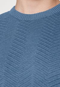 Pull en tricot à col rond, fabriqué en tissu bleu texturé présentant un subtil motif en chevrons. Doux et d'épaisseur moyenne.