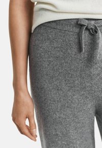 Woman wearing gray knit pants with a tied waistband and a white ribbed sweater, hand relaxed by side on a white background.