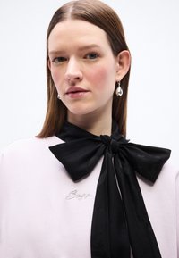 Νεαρή γυναίκα με ίσια καστανή τρίχα φοράει ασημένια σκουλαρίκια που κρέμονται, ένα ανοιχτό τοπ με μικρό λογότυπο σε γραφή και μια μεγάλη μαύρη κορδέλα δεμένη στον λαιμό της.