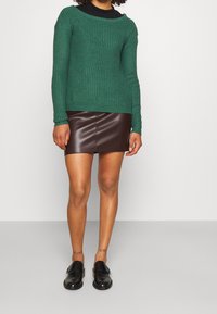 Dunkelgrüner, gerippter Strickpullover, kombiniert mit einem braunen Minirock aus Kunstleder. Schwarze Slipper runden das Outfit ab.