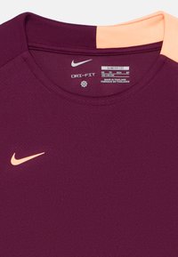 Lila Nike Dri-FIT póló kerek nyakkal, világos narancssárga vállpanellel díszítve. Vékony szabású, texturált szövetszövettel.