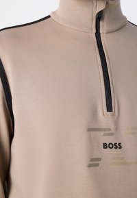 Beige zip-up top s černým zipem a pruhy na ramene, se textem "BOSS" a abstraktním horizontálním liniovým vzorem na hrudi.